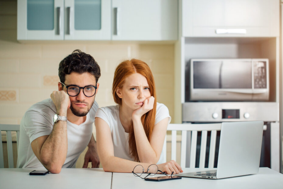 stressed couple sitting at the kitchen table with a laptop, worrying about money