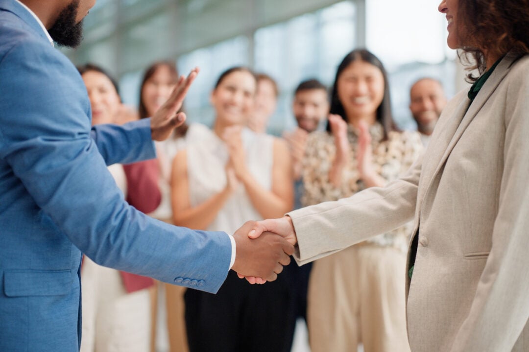 Two people in business attire shaking hands in the foreground. In the background, a diverse group of people stands clapping and smiling, suggesting a celebratory or successful event in a modern office setting.