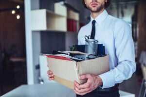 Man in a dress shirt and tie holding a box of his belongings after he was fired at work