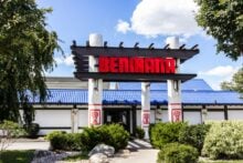 Front entrance of a Benihana restaurant, featuring a bold red sign on a white structure with decorative columns, surrounded by greenery and a bright blue roof under a partly cloudy sky.