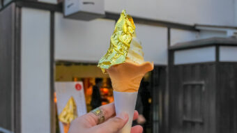A hand holding an ice cream cone with a swirl of vanilla ice cream, topped with a sheet of gold foil. The background shows a blurred exterior of a building.