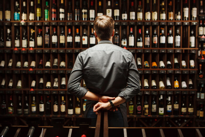 Bartender at wine cellar full of bottles with exquisite alcohol drinks that have various sweet and sour tastes and dates of manufacture on large wooden shelves.