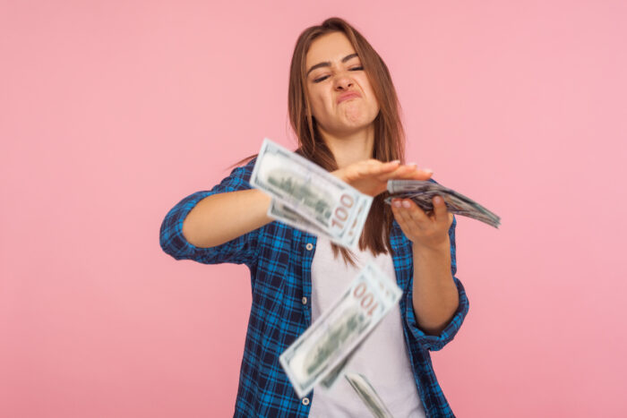 Portrait of wasteful rich girl in checkered shirt scattering dollars with arrogant grimace, boasting wealthy life, concept of careless money spending. indoor studio shot isolated on pink background