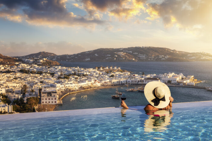 A tourist woman with a glas of wine in a swimming pool enjoys the view over the town of Mykonos island, Greece, during summer sunset time