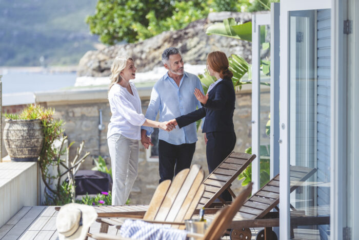 Real estate agent showing a mature couple a new house. The house is contemporary. All are happy and smiling and shaking hands. The couple are casually dressed and the agent is in a suit. Waterfront can be seen in the background