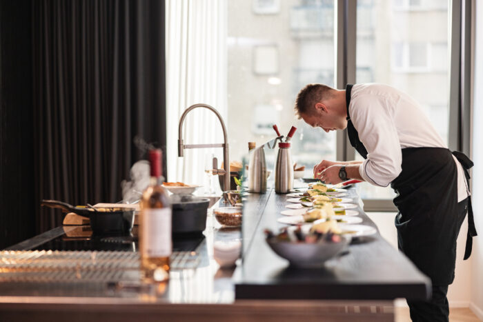 A young Caucasian male professional chef in an apron and uniform preparing food for the fancy bachelorette party. He is located in a modern kitchen with a view of the city. The chef is focused and precise while plating the food.