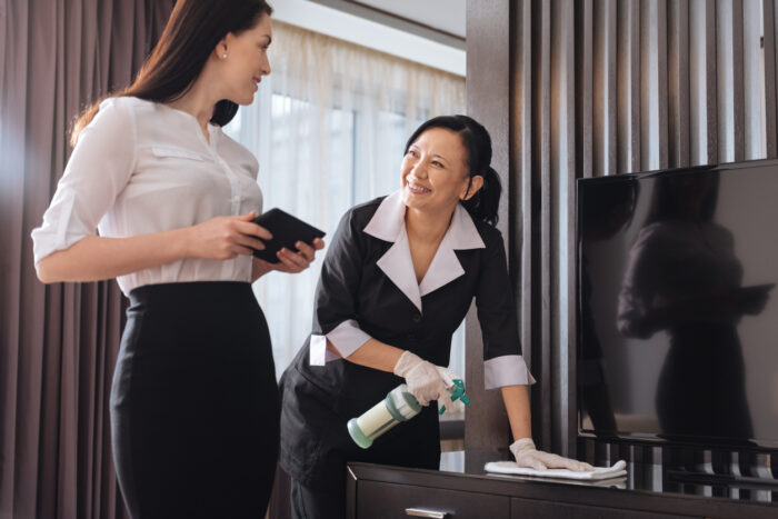 Hotel service. Joyful nice positive businesswoman standing near a hotel maid and talking to her while holding a tablet