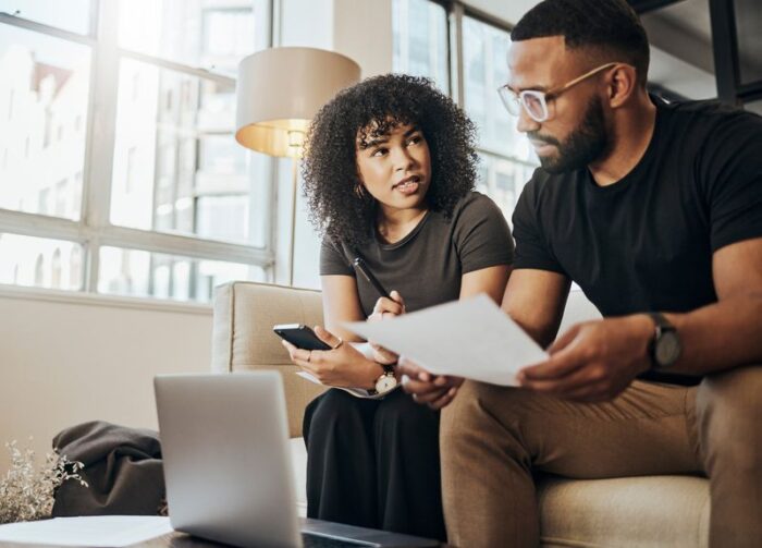 Finance, tax and couple talking about insurance, savings and information on a laptop with paper. Man and woman on the sofa of their house for ecommerce, banking and investing with technology