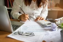 A person in a white shirt writes on a document at a cluttered desk, with a laptop, calculator, crumpled paper, eyeglasses, and a smartphone nearby. Sunlight falls on the workspace, suggesting a busy work session.