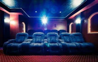 A dimly lit home theater with rows of plush, blue reclining seats facing a bright projector screen. The room has red walls, arched doorways with glowing outlines, and a star-like pattern on the carpeted floor.