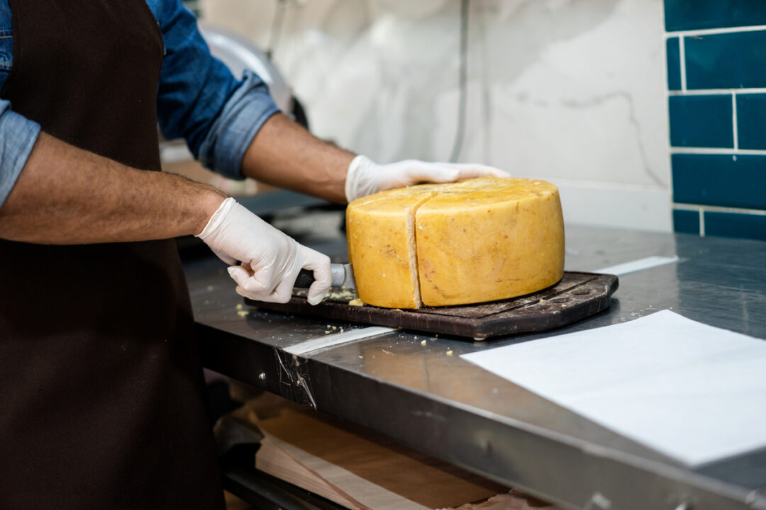 Close-up of salesman cutting aged cheese on grocery shop counter. Male delicatessen employee hands wearing gloves slicing large cheese wheel.