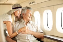 Older couple smiling and looking out the window of a private plane. The man is seated, wearing a light shirt, while the woman leans over him, wearing a straw hat and white top. Sunlight streams through the windows, creating a warm atmosphere.