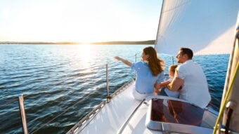 A family of three is sitting on a sailboat, enjoying a sunny day. They are gazing at the water as the sunlight glistens on the waves. The woman is pointing out towards the horizon while the man embraces the child.
