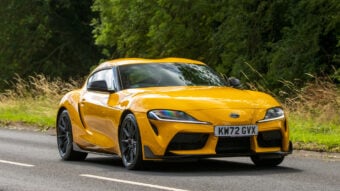 Yellow sports car driving on a road with a lush, green forest in the background. The car has sleek lines and modern detailing, and the license plate reads "KW72 GVX.