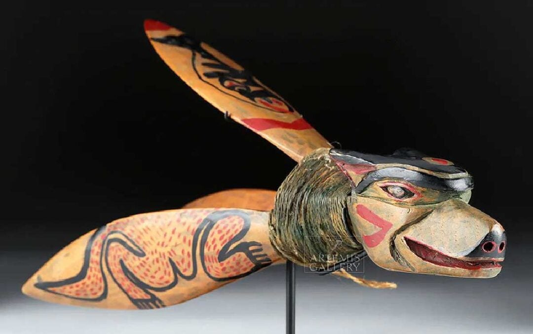 A wooden mask shaped like a stylized animal head with large, painted wings, featuring bold red, black, and cream designs, displayed on a stand against a dark background.