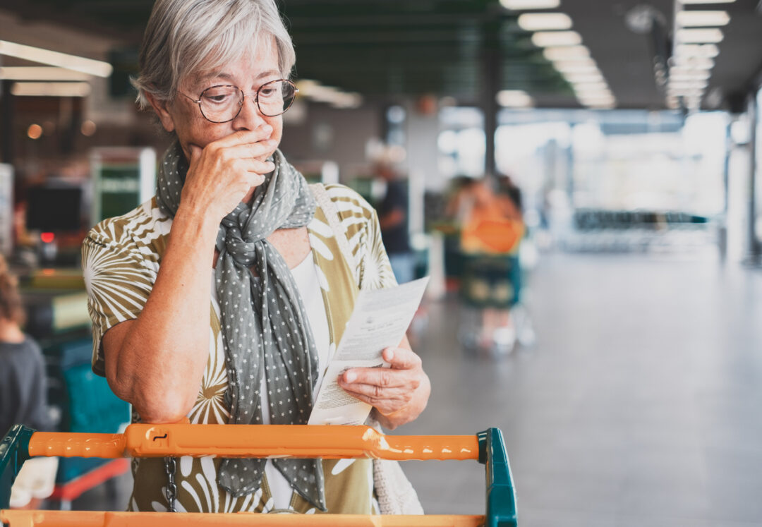 Senior woman in the supermarket checks her grocery receipt looking worried about rising costs - elderly lady pushing shopping cart, consumerism concept, rising prices, inflation