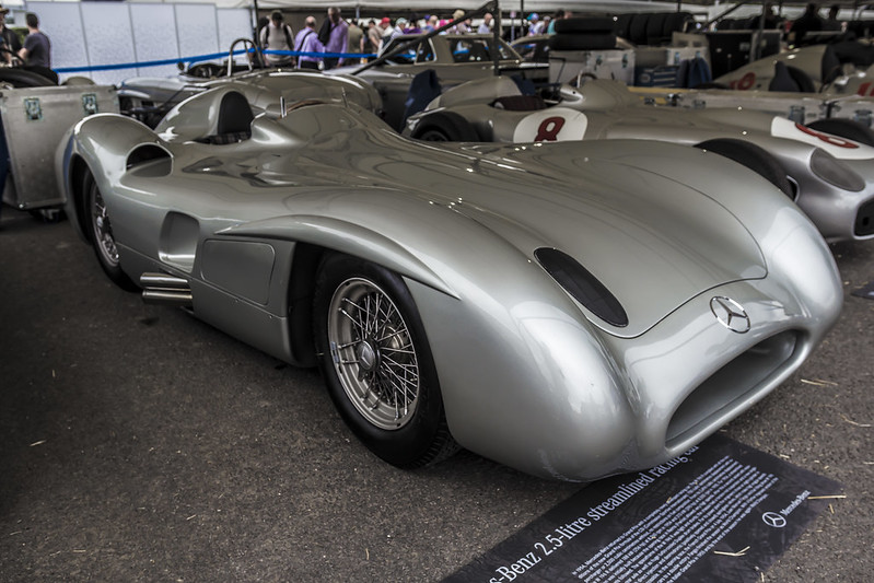 A sleek, silver vintage Mercedes-Benz race car with a streamlined body is displayed in an exhibition setting. The car features wire-spoke wheels and a classic emblem on the front. Other vintage cars are visible in the background.