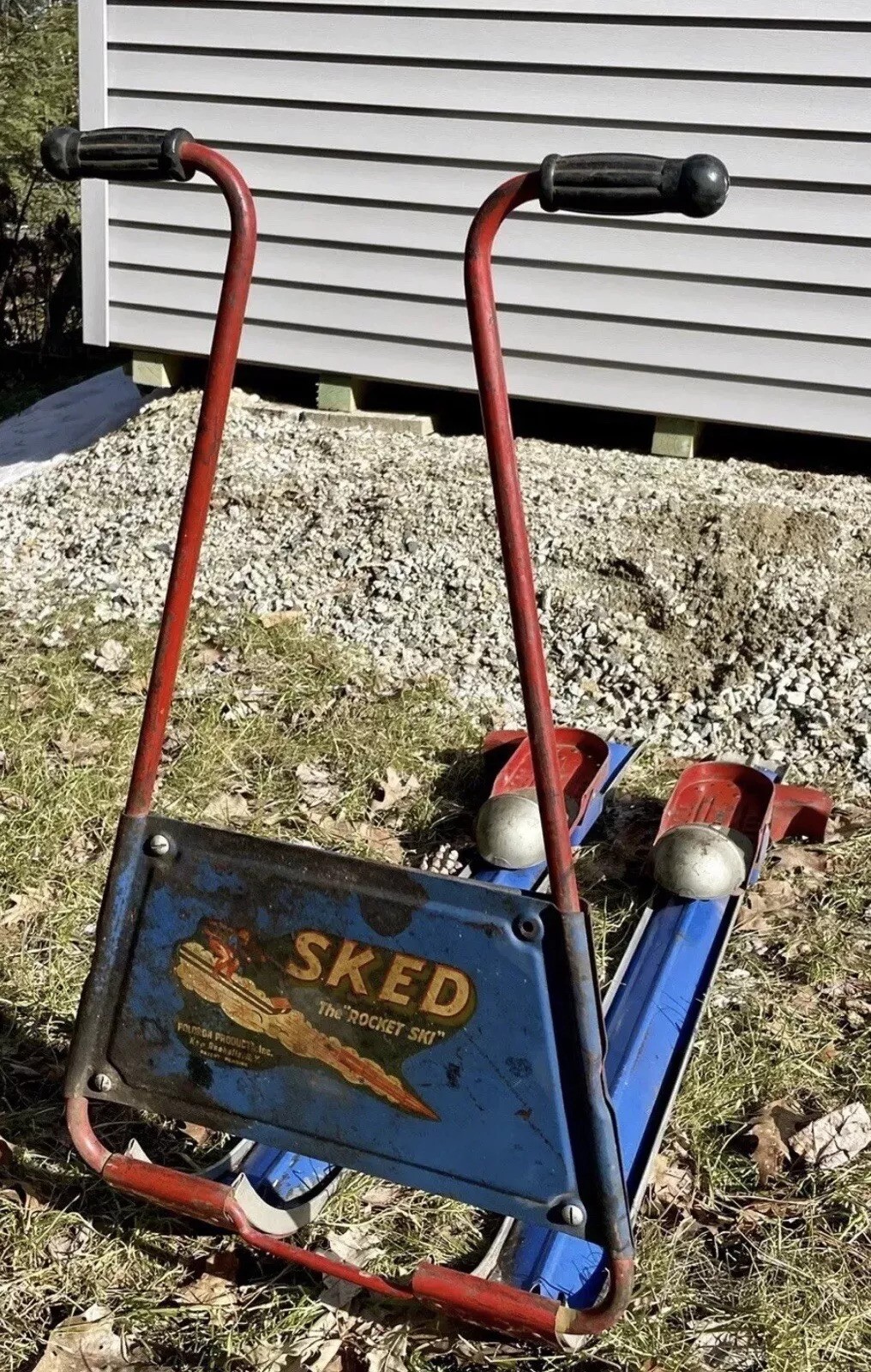 A vintage red and blue metal scooter-like device named "Sked" with the tagline "The Rocket Ski" on a blue panel. It has T-shaped handlebars and rests on a grassy area with gravel and a gray building in the background.