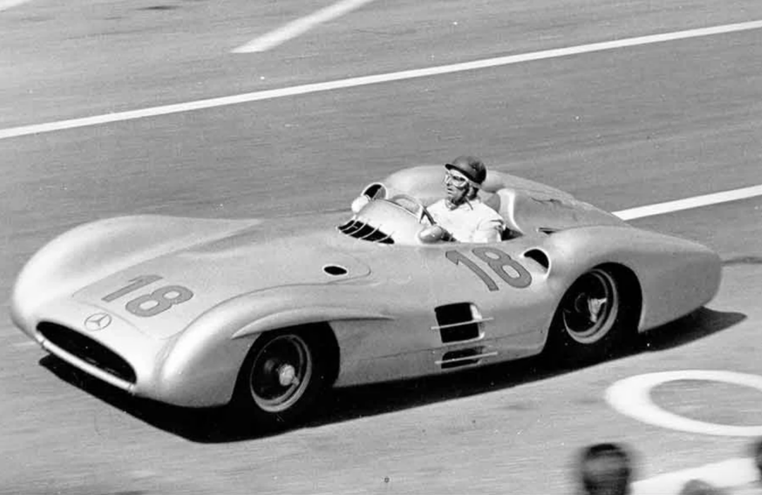 A vintage race car, marked with the number 18, is speeding down a track. The driver wears a helmet and is seated in an open cockpit, showcasing the sleek and aerodynamic design typical of mid-20th century racing vehicles.