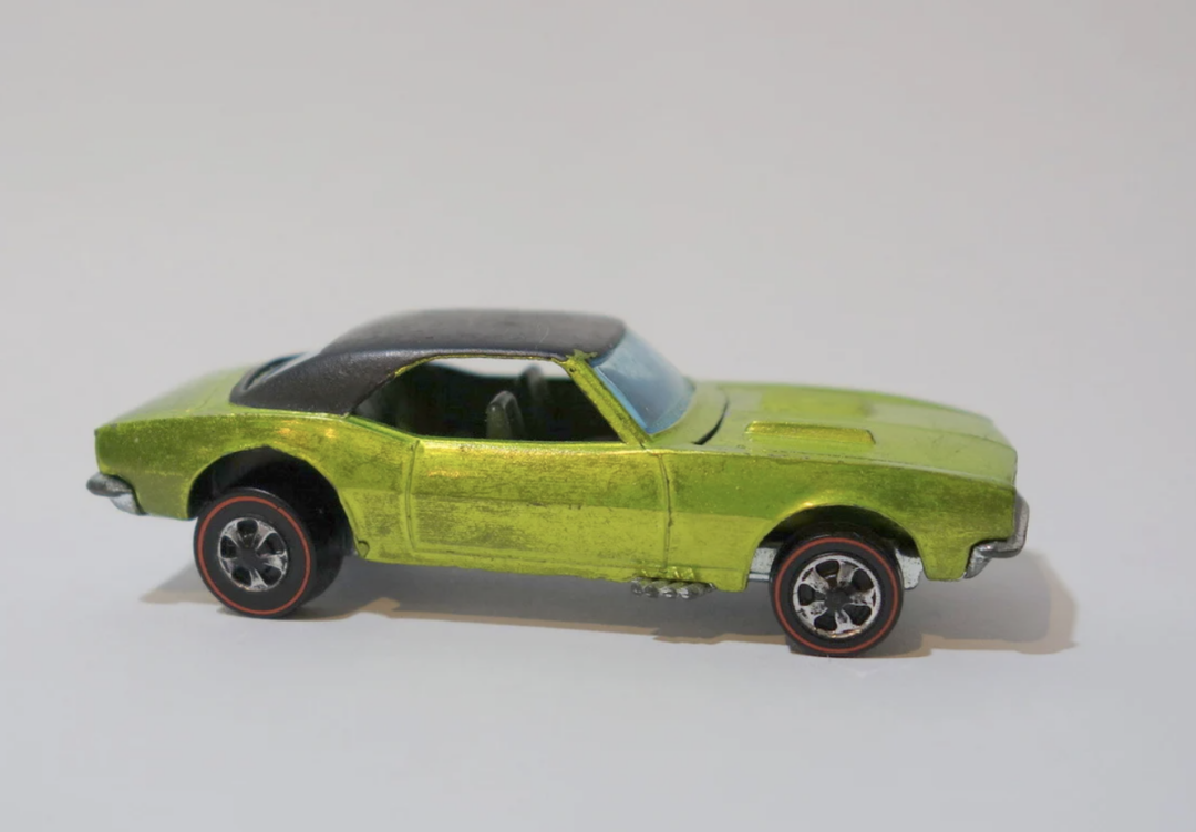 A small Hot Wheels toy car with a shiny green body and a black roof, viewed from the side. It features red-lined black wheels and silver detailing, set against a plain white background.