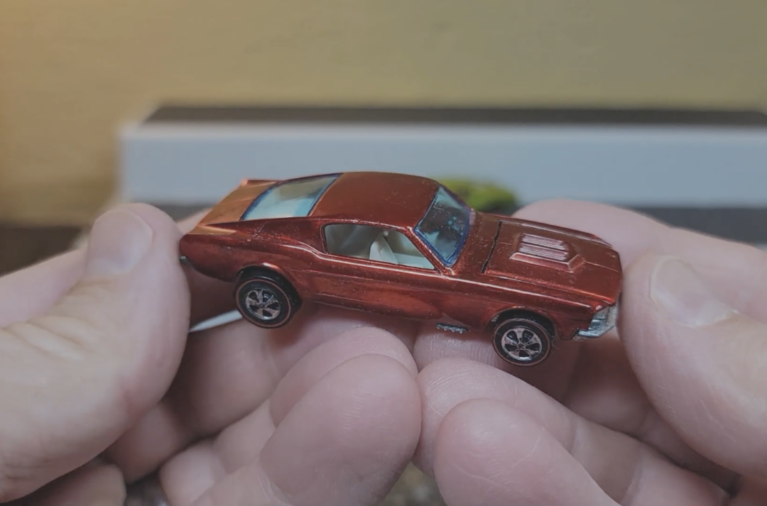 A person's hands holding a small, vintage Hot Wheels toy car with a boxy shape and detailed features, set against a blurred background.