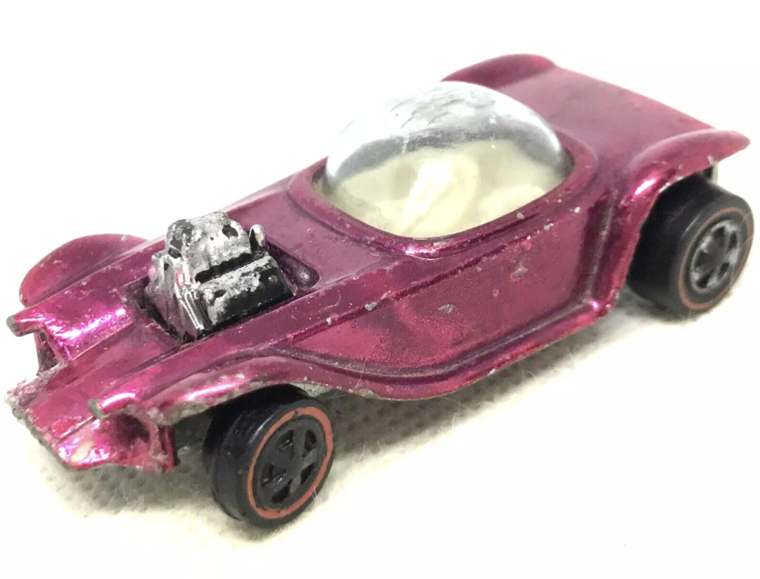 A vintage Hot Wheels toy car with a shiny magenta finish, featuring an exposed engine and a transparent domed windshield. The car's body shows some charming wear and tear, complemented by black wheels with striking red trim.