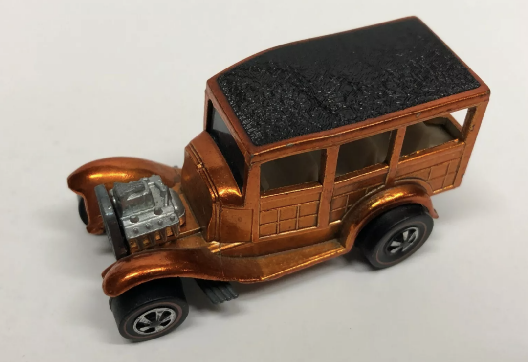 A small toy car resembling a vintage model, with a bronze body and black roof, evokes the charm of Hot Wheels. It features four wheels and an exposed engine. With its classic rectangular shape, this miniature marvel is displayed on a plain surface.