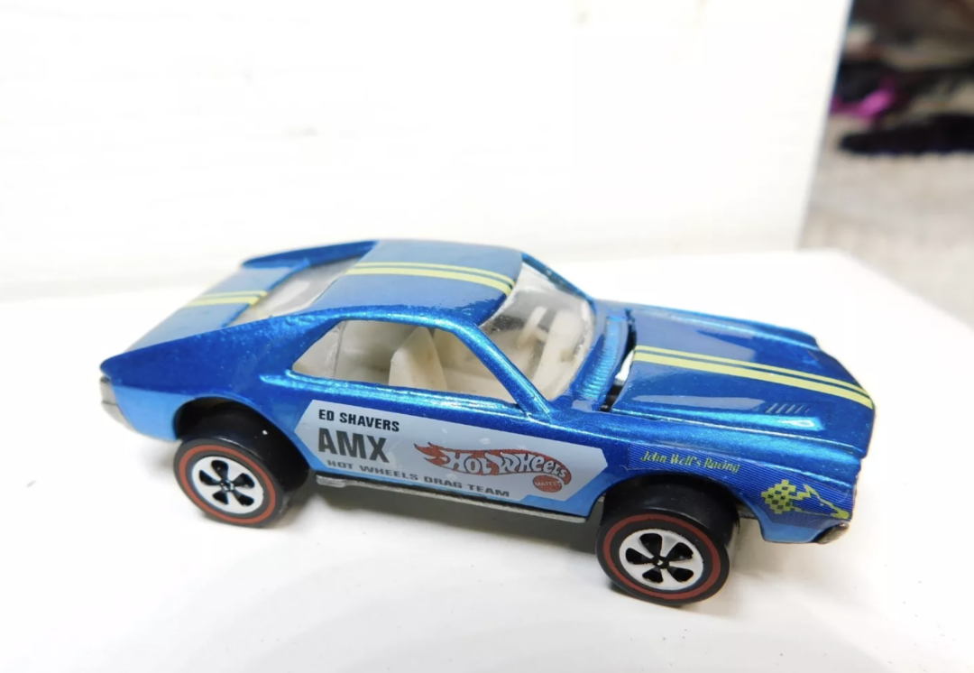 A close-up of a blue Hot Wheels toy car with black and yellow stripes. The sides display logos and text, including "ED SHAVERS," "AMX," and "Hot Wheels Drag Team." It features red-lined wheels and a shiny finish.