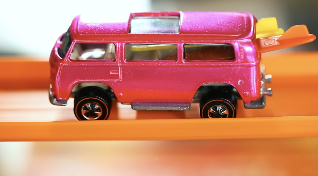 A close-up of a small toy van painted in metallic pink, styled with surfboards on the back. It's positioned on an orange Hot Wheels track, suggesting motion or play. The van has black wheels with silver rims and detailed windows, capturing the thrill of tiny adventures.