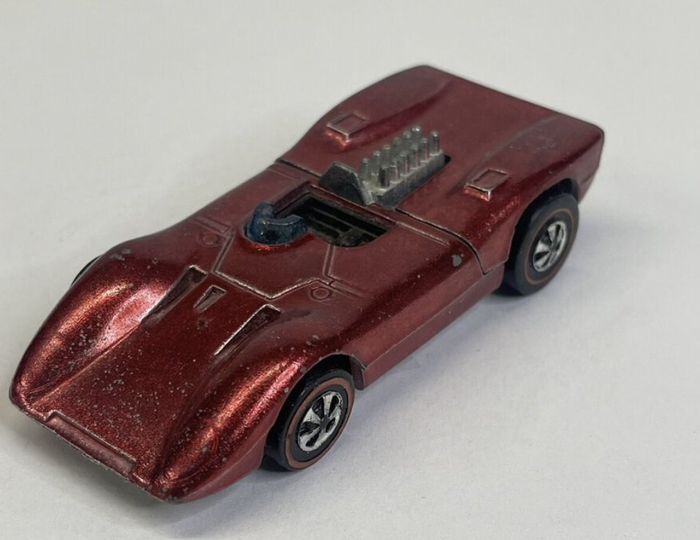 A vintage, red die-cast Hot Wheels toy car with a sleek, aerodynamic design. The car features prominent front fenders, a visible engine detail, and black wheels with white rims, set against a plain white background.