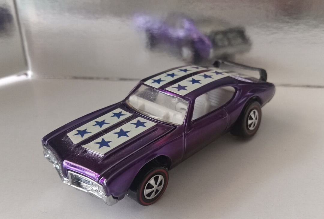 A purple Hot Wheels toy car, adorned with white stripes and blue stars on the roof, sits on a reflective surface. Its red-striped wheels and rear spoiler add flair, with its shimmering reflection visible in the background.