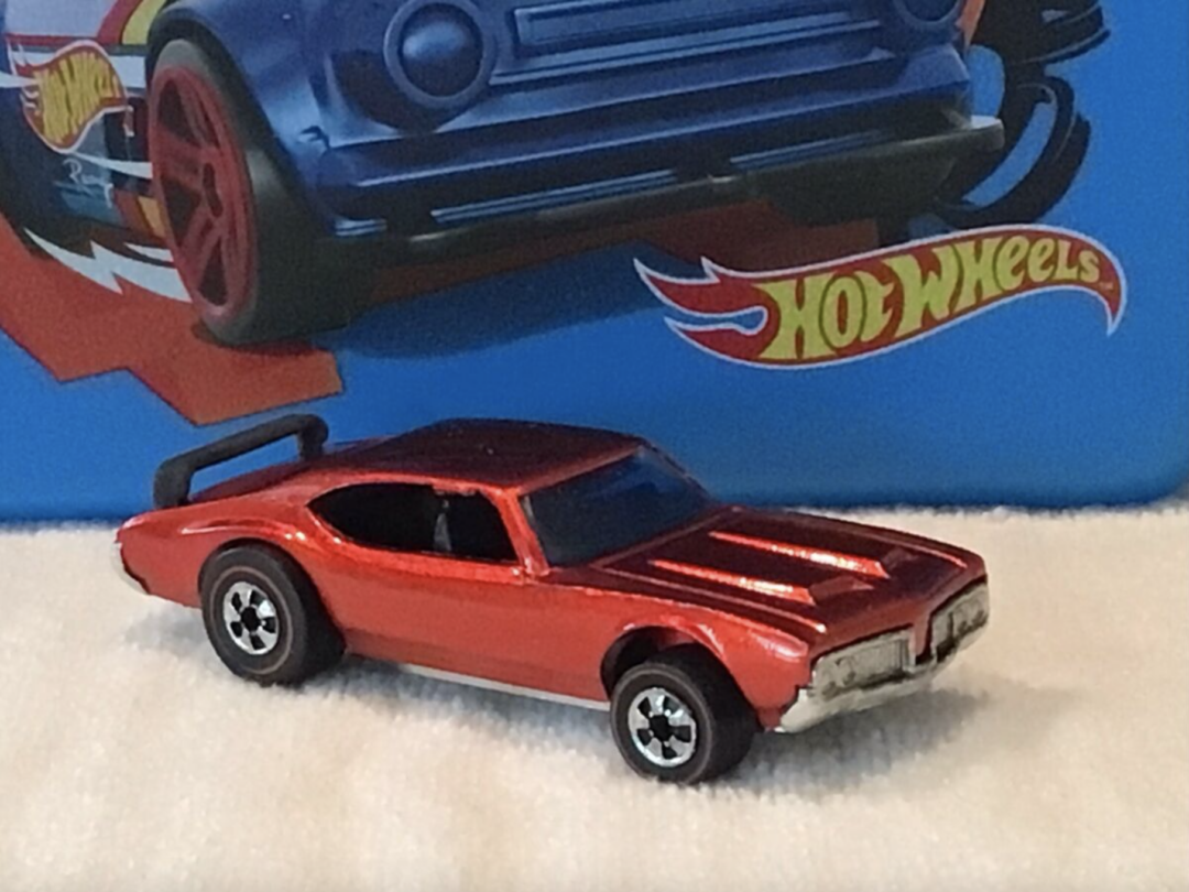 A red toy car with black details is sitting on a white surface. In the background, the iconic Hot Wheels branding adorns a blue box, which also features a partially visible image of another toy car.