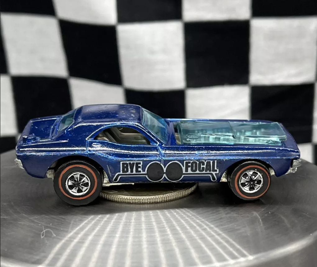 A blue Hot Wheels toy car with "BYE FOCAL" emblazoned on its side sits on a round platform. The backdrop showcases a black and white checkered pattern, accentuating the car's silver and red details.