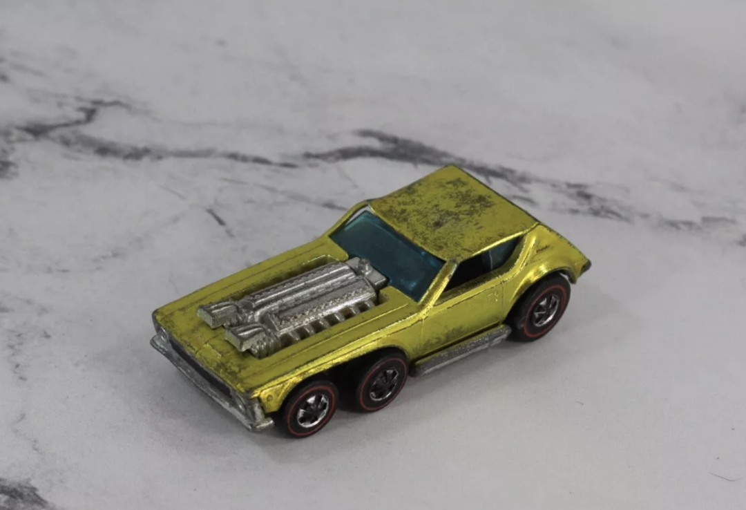 A close-up of a well-worn yellow Hot Wheels car with an exposed silver engine on a gray marble surface. The toy boasts a sporty design and red-lined wheels, bearing the marks of age and countless adventures.