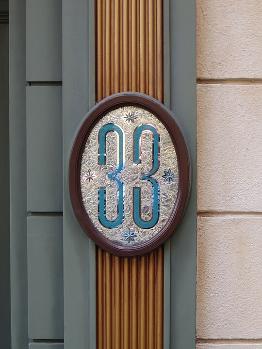 An ornate oval sign displaying the number 33 with decorative patterns and stars, mounted on a wall with vertical wood and green trim detailing.