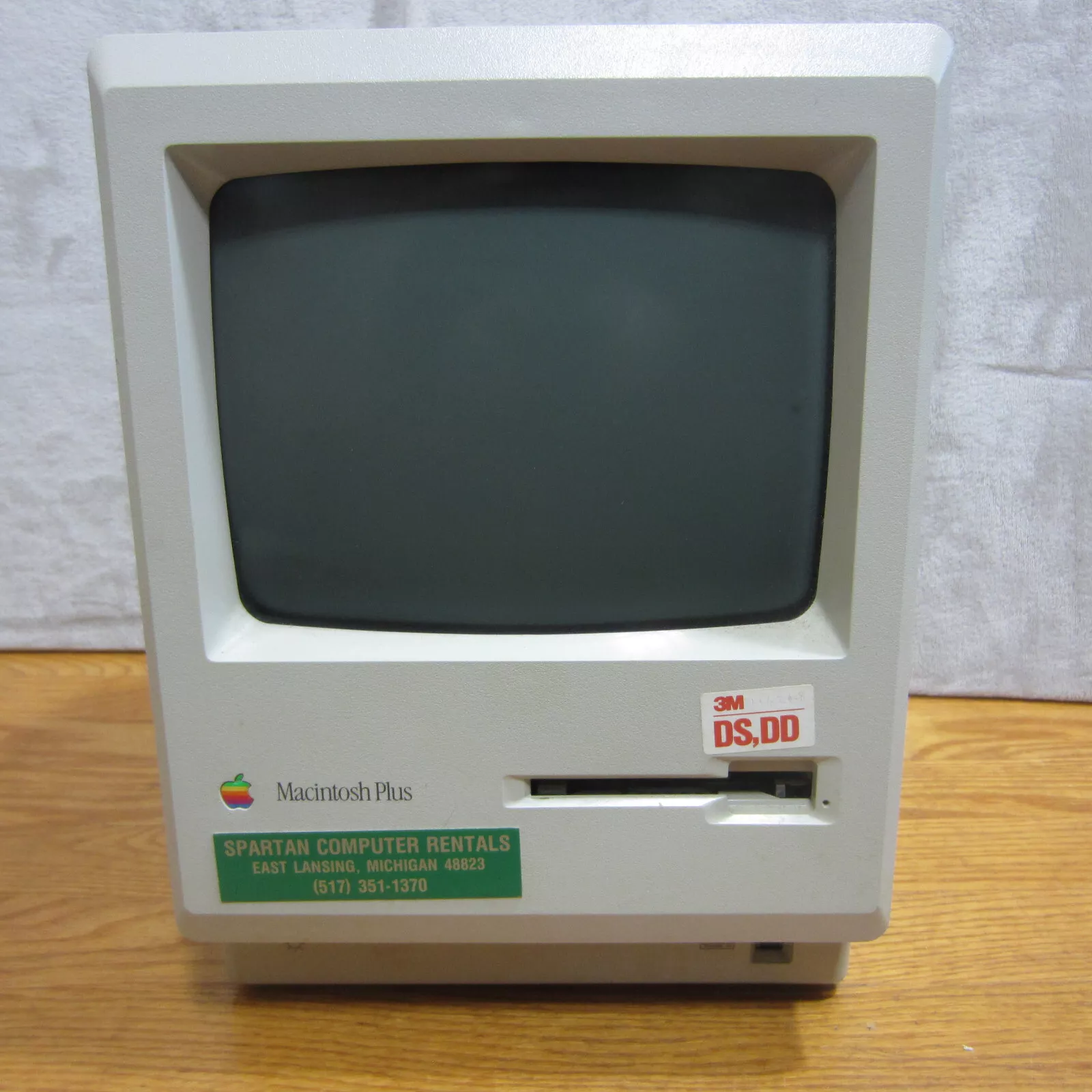 A vintage Macintosh Plus computer with a floppy disk drive, featuring a beige exterior and a green label from Spartan Computer Rentals, East Lansing, Michigan, on a wooden surface.