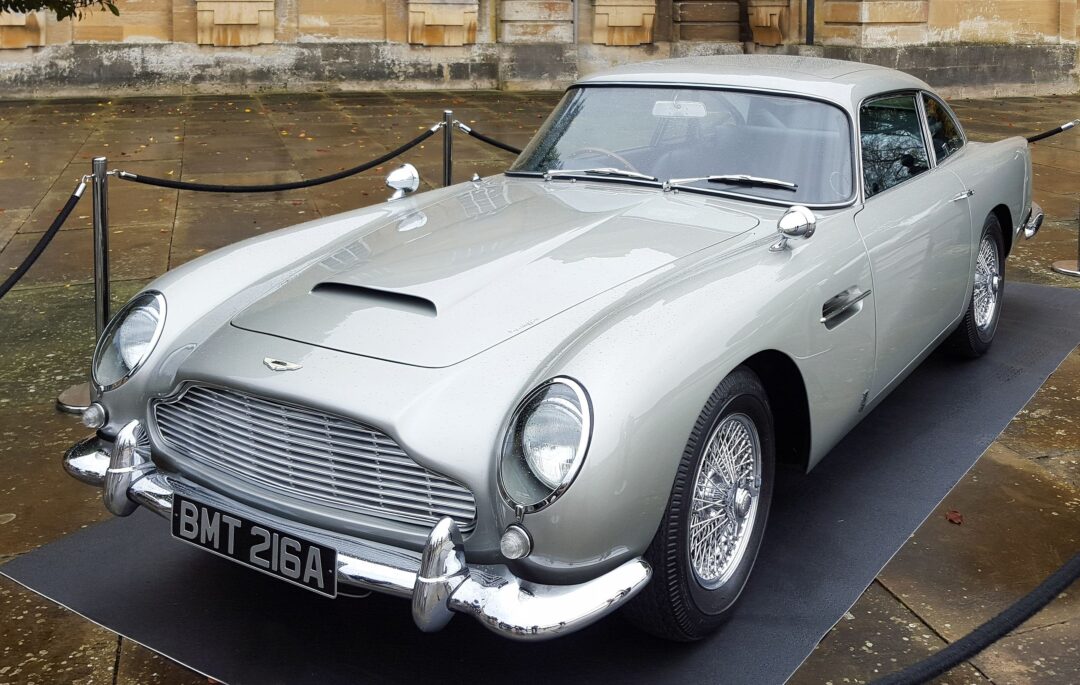 A classic silver vintage car with chrome detailing and a "BMT 216A" license plate is displayed outdoors on a black platform, surrounded by ropes and a stone pavement.