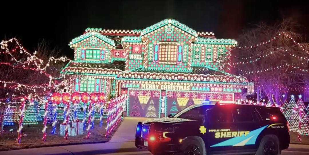 A house adorned with elaborate Christmas lights features a "Merry Christmas" sign and various colorful displays. In front, a sheriff's vehicle with lights on is parked in the driveway at night.