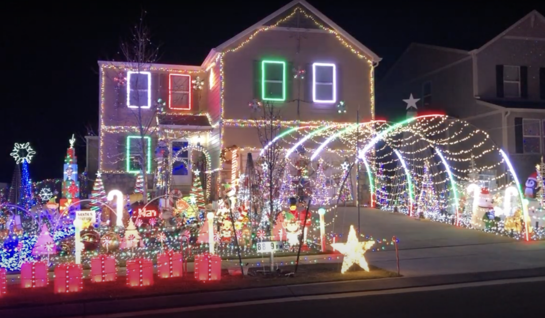 A house is brightly decorated with colorful Christmas lights and ornaments. The display includes a tunnel of lights over the driveway, various festive figures in the yard, and illuminated stars and presents, creating a vibrant holiday scene.