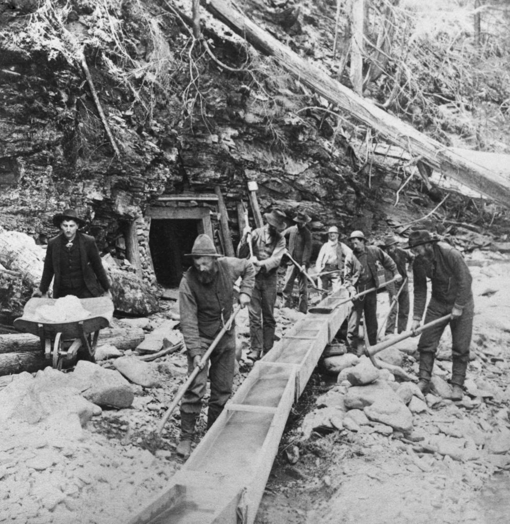 A group of men in vintage clothing work at a mining site, using shovels and a sluice box. They are surrounded by rocky terrain and trees, with a tunnel entrance visible in the background. One man holds a wheelbarrow.