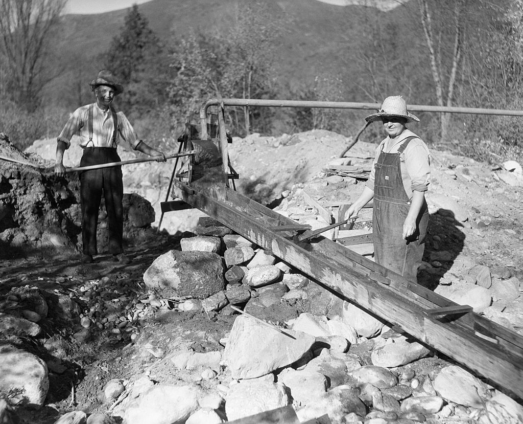 Two people in overalls and hats operate a sluice box in a rocky outdoor setting, likely during a gold mining activity. One person holds a shovel, while the other stands near the sluice. Trees and mountains are visible in the background.