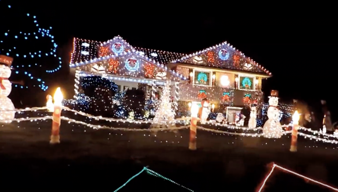 A house decorated with Christmas lights at night. The roof and windows are outlined with colorful lights, and there are illuminated snowmen and candy cane decorations on the lawn. String lights create a festive ambiance.