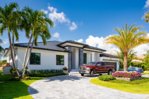 A modern, single-story house with a light gray exterior, large windows, and a dark roof. A red pickup truck is parked in the curved driveway. Palm trees and vibrant landscaping surround the home under a clear blue sky.