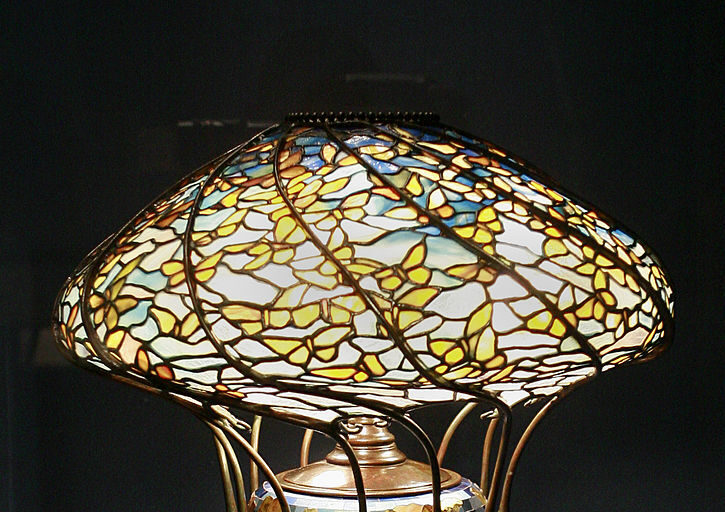 This elegant, expensive Tiffany-style lamp boasts an ornate stained glass design with a floral pattern and a wide, mushroom-like shade. The tall, vase-shaped base is adorned with colorful mosaic designs, emitting a warm glow that highlights its intricate metalwork and craftsmanship.