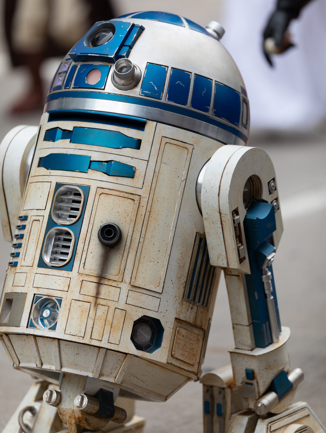 A close-up of a detailed replica of a blue and white droid, resembling R2-D2 from Star Wars. The droid's design includes various panels and lights, and it appears to be outdoors. Blurred figures are visible in the background.