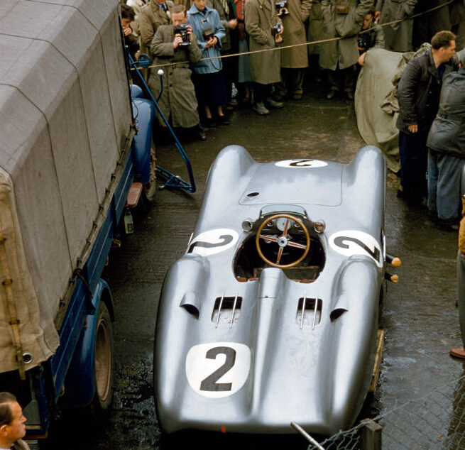 A vintage race car with the number 2 on it is parked near a crowd of onlookers. The car is positioned next to a truck and surrounded by spectators in coats. A fenced area is partially visible in the foreground.