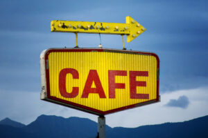 A vintage sign with large red letters spelling "CAFE" on a yellow background is topped with a weathered, yellow arrow pointing right. The sky is overcast, and mountains are faintly visible in the background.