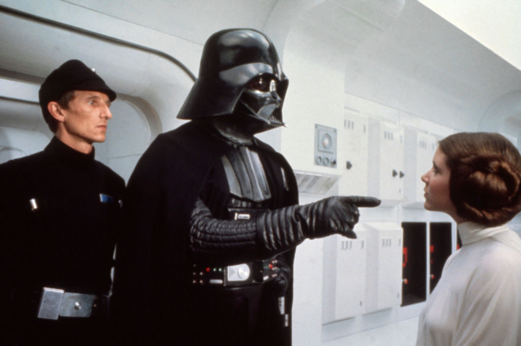 A person in black armor and a black cape is pointing at another person wearing a white robe with a braided hairstyle. A third person in a dark uniform and cap stands nearby in a futuristic interior with control panels.