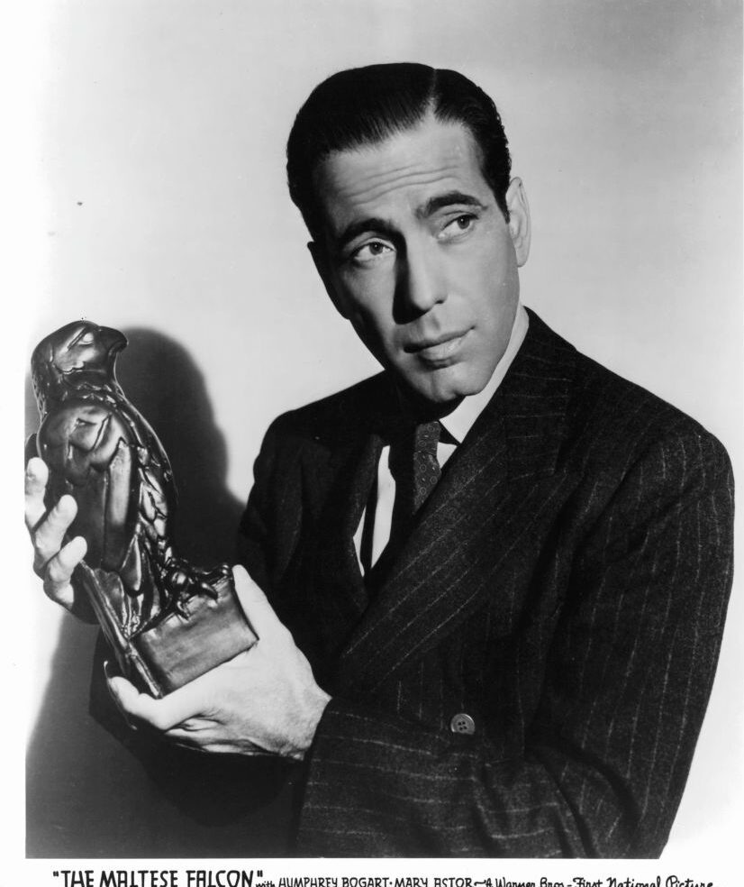 A black and white photo of a man in a suit holding a falcon statuette. He looks to the side with a serious expression. The image is titled "The Maltese Falcon.