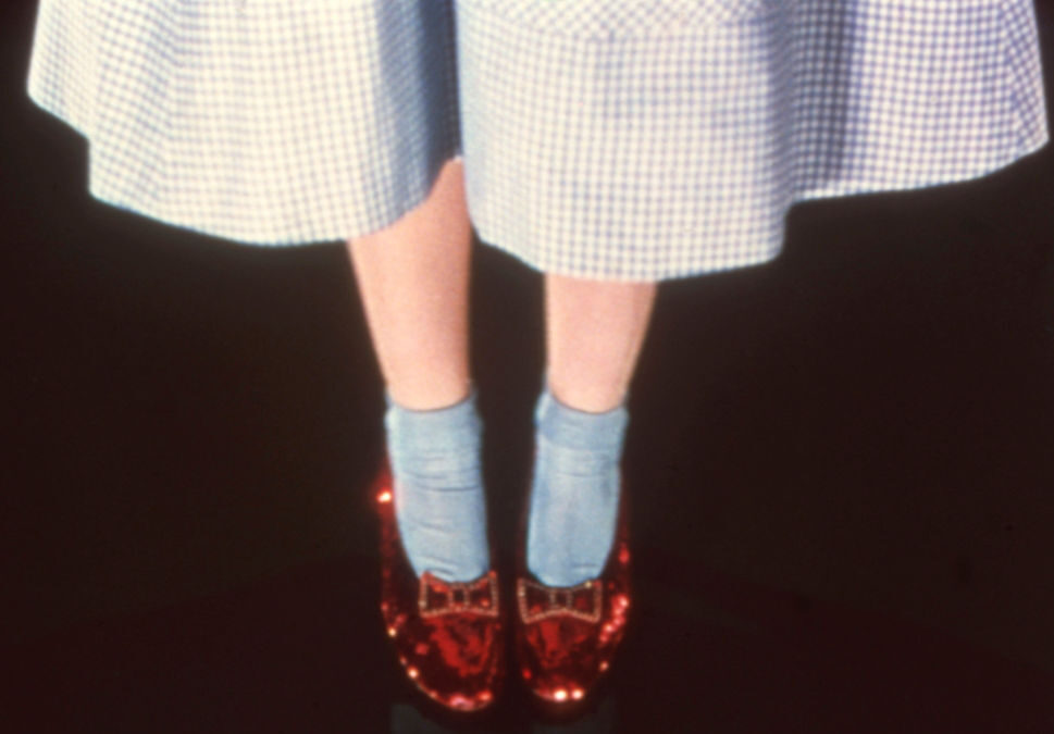 A person wearing blue socks and shiny red shoes with bows stands against a dark background. They are dressed in a knee-length, checkered skirt.